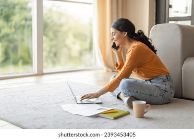 Portrait Of Young Indian Woman Using Laptop And Talking On Cellphone At Home, Smiling Eastern Female Working Or Study Remotely With Computer While Sitting On Floor In Living Room, Copy Space - Powered by Shutterstock