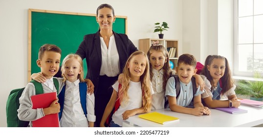 Portrait Of Young Intelligent Female Teacher With Her Little Pupils Of Primary Class. Smiling Boys And Girls 6-7 Years Old Together With Their Teacher Smile At Camera In School Classroom. Banner.