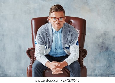 Portrait Of A Young Intelligent Man Stares Into The Camera, Good View, Small Unshaven, Charismatic, Wearing Glasses Rimmed With Brown, Blue Shirt, Gray Sweater, Sitting On A Brown Leather Chair