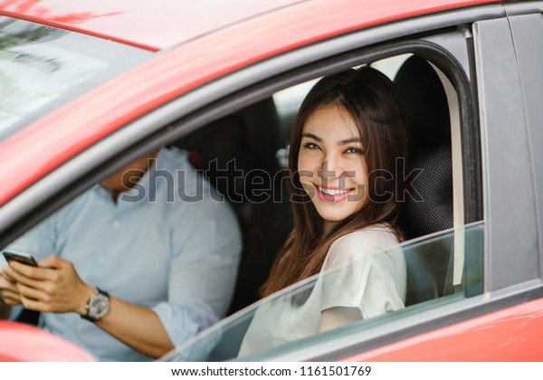 Portrait of\
a young Japanese Asian woman leaning out the window of a red car\
window and smiling happily. She is being driven to her destination\
in a ride she booked on a ride hailing app.\
