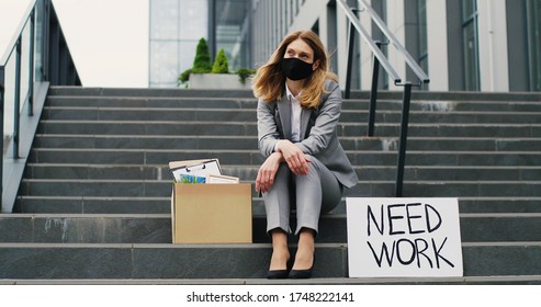 Portrait Of Young Jobless Caucasian Woman In Mask Sitting On Stairs Outdoor With Carton Poster Need Work. Unemployment After Pandemic Lockdown. Female Fired Office Worker Looking At Stuff In Box.