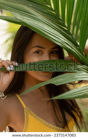 Portrait of young joyful beautiful Asian woman with long dark hair, wearing yellow top, looking through leaf of palm tree, hiding, posing, having fun in tropical resort. Beauty, travelling. Vertical.