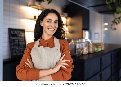 Portrait of young joyful successful Latin American small business owner, slim boss smiling and looking at camera with crossed arms, businesswoman at entrance to cafe restaurant, inviting visitors. - Powered by Shutterstock