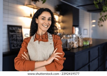 Portrait of young joyful successful Latin American small business owner, slim boss smiling and looking at camera with crossed arms, businesswoman at entrance to cafe restaurant, inviting visitors.