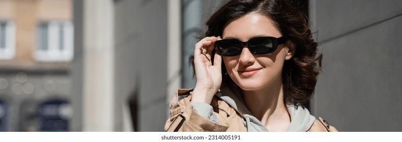 portrait of young and joyful woman with wavy brunette hair, wearing beige trench coat and grey hoodie, adjusting dark stylish sunglasses and looking away on urban street, banner - Powered by Shutterstock