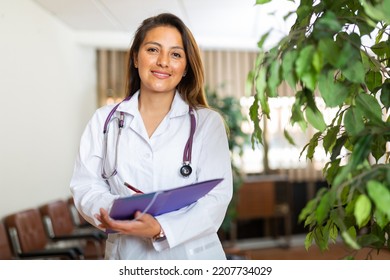 Portrait of young latin american female doctor wearing white coat standing in clinic office, filling out medical form at clipboard. - Shutterstock ID 2207734029