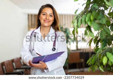 Portrait of young latin american female doctor wearing white coat standing in clinic office, filling out medical form at clipboard.