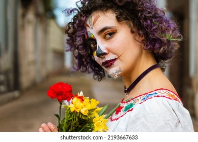 portrait of young latin caucasian argentinian woman, holding flowers with her hands standing outdoors in the cemetery with La Calavera Catrina makeup, looking at the camera in profile.