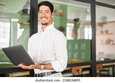 Portrait Of Young Latin Guy Standing And Holding Laptop In Contemporary Office Space, Male White Collar Worker Looks At Camera, Posing Indoors With Glass Loft Style Partition On Background
