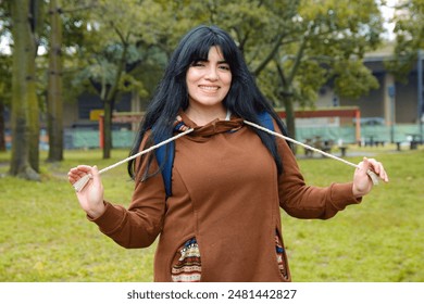 Portrait of young Latin tourist woman smiling and playing with strips of hood of her brown sweater, walking through park, she is standing posing for camera, she is Venezuelan immigrant woman. - Powered by Shutterstock