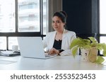 Portrait of young latina woman in the office sitting working with the laptop she is smiling and looking at the camera and smiling, she is typing making reports and marketing strategies for the company