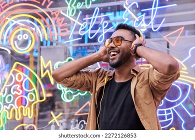 Portrait of young latino boy with neon light reflection on his face listening to music. Boy looking to the side with hands on helmets. Hipster millennial with beard and sunglasses. Horizontal image.
