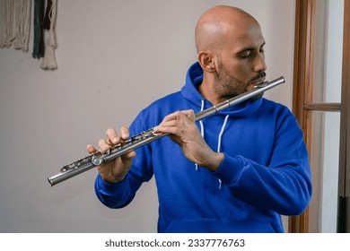 portrait of young latino man in blue, with beard and bald head standing at home playing flute with inspiration