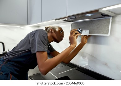 Portrait Of Young Lovely African Ethnicity Repairwoman With Flashlight, Changing Kitchen Exhaust Filters. Handywoman In Modern Light Kitchen. Authenticity In Business Concept.