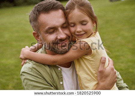 Portrait of young loving father and his cute little girl keeping eyes closed and hugging while spending time outddors, sitting on a green grass