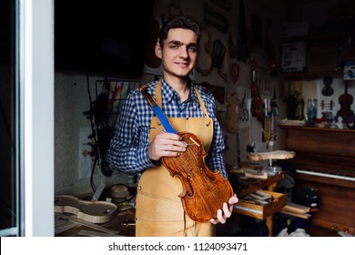 portrait of a young luthier with a handcrafted violin in his workshop with the tools and the woods on the background. Master artist poses in his home workshop. Holds violin-workpiece. - Powered by Shutterstock