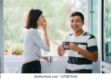 A portrait of a young Malay Asian couple enjoying a hot drink and talking to one another fondly during the day by the window balcony. They are both laughing naturally as they share a joke together.  - Powered by Shutterstock