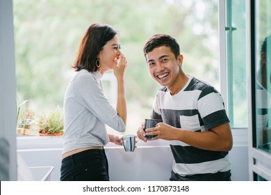 A portrait of a young Malay Asian couple enjoying a hot drink and talking to one another fondly during the day by the window balcony. They are both laughing naturally as they share a joke together.  - Powered by Shutterstock