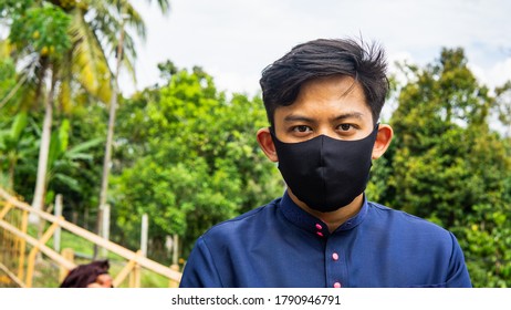 A portrait of a young Malay man with traditional Baju Melayu cloth wearing a black fabric face mask with green nature background. Protection step to avoid spreading viruses and diseases.