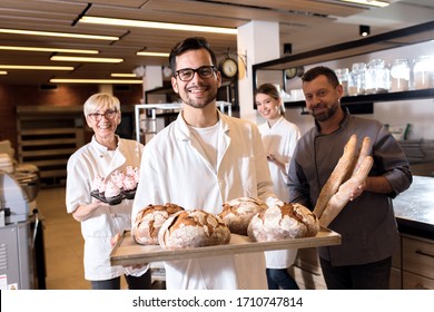 Portrait Of Young Male Baker Holding Bread In His Hands At Bakery With His Multi Generation Colleagues In Backgrounds.