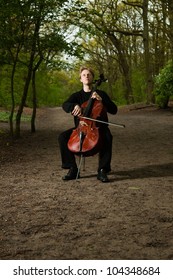 Portrait Of Young Male Cello Player In Green Spring Forest. Blond Hair. Playing Classic Instrument. Dressed In Black. Artistic Looking.