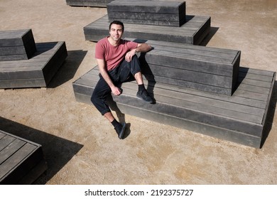 Portrait Of Young Male Dancer Sitting On Geometrical Benches In A London Park. He Is Smiling And Looking At Camera.