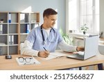 Portrait of a young male doctor or nurse in blue medical uniform sitting at the desk and working in office. Man physician wearing stethoscope in clinic using laptop typing and writing in notepad.