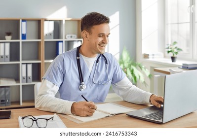 Portrait of a young male doctor or nurse in blue medical uniform sitting at the desk and working in office. Man physician wearing stethoscope in clinic using laptop typing and writing in notepad. - Powered by Shutterstock