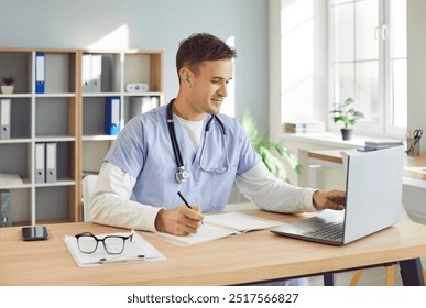 Portrait of a young male doctor or nurse in blue medical uniform sitting at the desk and working in office. Man physician wearing stethoscope in clinic using laptop typing and writing in notepad. - Powered by Shutterstock