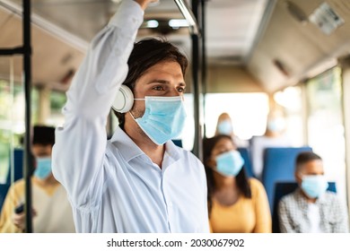 Portrait of young male passenger wearing medical face mask standing traveling on public transit, listening to music in wireless headset and looking away at window, thinking, going to work by bus
