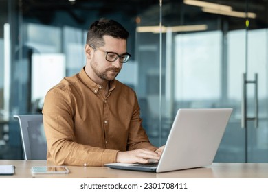 Portrait of a young male programmer, in a brown shirt and glasses who is concentrating on working on a laptop, while sitting at a desk in the office. - Powered by Shutterstock