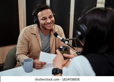 Portrait of young male radio host going live on air, talking with female guest, holding a script paper while sitting in studio
