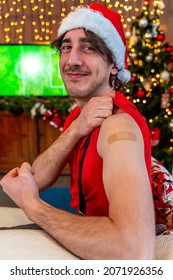 Portrait Of A Young Male Smiling After Getting A Vaccine - Man Showing Her Arm With Bandaid During A Christmas Party Dressing Red Cap