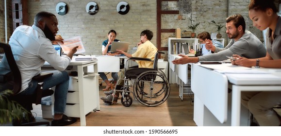 Portrait of young male worker in wheelchair talking to female colleague while presenting ideas using laptop in co-working space. Creative team at work. Disability and handicap concept. Horizontal shot