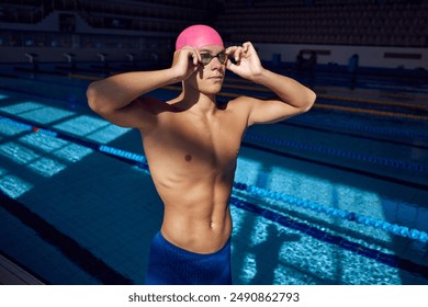 Portrait of young man with athletic body, swimmer wearing goggles and pink cap, posing against blurred background of pool. Concept of aquatic sport, preparation to competition, energy. - Powered by Shutterstock