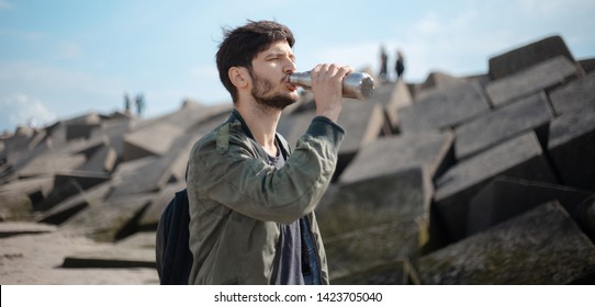 Portrait Of Young Man With Backpack, Using Reusable Steel Thermo Water Bottle, Outdoor On Background Of Square Stones.