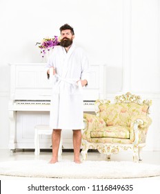 Portrait Of A Young Man In A Bathrobe Standing Isolated In White Room With Piano. Bearded Guy Tightening His Robe. Sleepy Hipster In His Apartment.