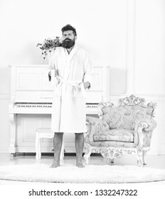 Portrait Of A Young Man In A Bathrobe Standing Isolated In White Room With Piano. Bearded Guy Tightening His Robe. Sleepy Hipster In His Apartment.