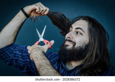 Portrait Of A Young Man With A Beard And Long Hair Who, With A Scissor In His Hand, Wants To Change His Look And Wants To Cut His Hair, Concept To Illustrate The Desire To Change