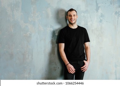 portrait of a young man in a black T-shirt leaned against a vintage gray wall. Copy space. Masculine model of bristles. Happy smiling.