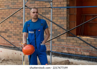 Portrait Of A Young Man In Blue Construction Overalls At A Construction Site. High Quality Photo