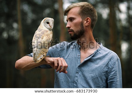 Portrait of young man in blue shirt with owl in forest. Owl sits on his hand and guy looks at the owl. Close-up.