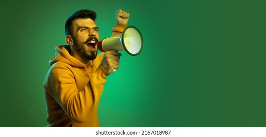 Portrait Of Young Man In Casual Cloth Shouting In Megaphone Isolated Over Green Studio Background. Motivational Speech. Concept Of Emotions, Facial Expression, Lifestyle, Fashion, Youth Culture