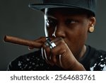 Portrait of a young man with a cool attitude smoking a cigar, wearing a baseball cap and rings, looking intensely at the camera against a dark background