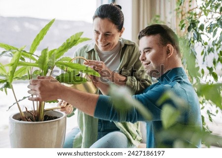 Portrait of young man with Down syndrome with his mother at home, taking care of plants. Concept of love and parenting disabled child.