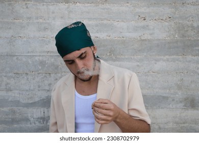 Portrait of a young man dressed in beige suit and green headscarf lights a cigarette, gray wall background. May 31 world day against smoking. Concept social problem. - Shutterstock ID 2308702979