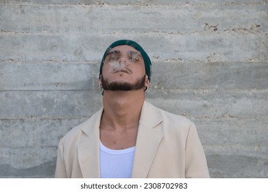 Portrait of a young man dressed in beige suit and green headscarf smoking a cigarette, gray wall background. May 31 world day against smoking. Concept social problem. - Shutterstock ID 2308702983