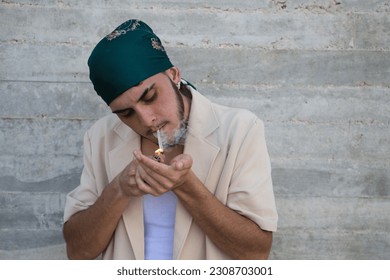 Portrait of a young man dressed in beige suit and green headscarf lights a cigarette, gray wall background. May 31 world day against smoking. Concept social problem. - Shutterstock ID 2308703001