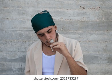 Portrait of a young man dressed in beige suit and green headscarf lights a cigarette, gray wall background. May 31 world day against smoking. Concept social problem. - Shutterstock ID 2311933297