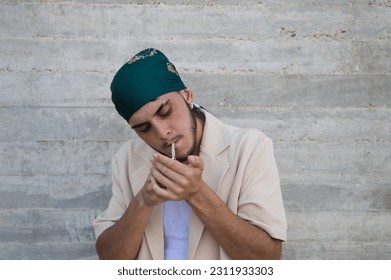Portrait of a young man dressed in beige suit and green headscarf lights a cigarette, gray wall background. May 31 world day against smoking. Concept social problem. - Shutterstock ID 2311933303